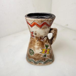 May include: A ceramic mug with a flared top and a single handle. The exterior features a beige base with a dark brown wash, decorated with hand-painted geometric patterns in orange, yellow, green, and red. The interior rim is painted black.