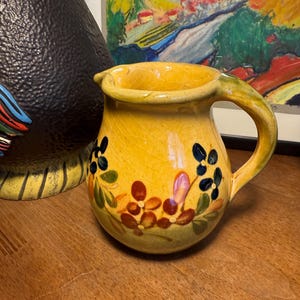 Può includere: Una brocca in ceramica gialla con un manico curvo e un beccuccio. La brocca è decorata con disegni floreali nei toni del rosso, blu e verde. La brocca è appoggiata su una superficie di legno.