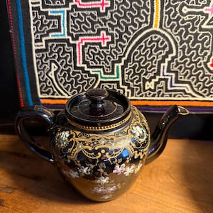 May include: A black ceramic teapot with a lid, handle, and spout. The teapot is decorated with gold floral patterns and small, colorful flowers. The background features a patterned textile.