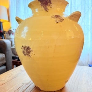 May include: A large, yellow ceramic vase with two side handles and a wide opening. The vase has a distressed finish with brown spots, giving it a rustic appearance. The vase is sitting on a wooden table.