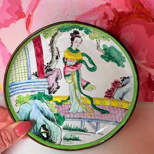 May include: A small, round, hand-painted decorative plate with a detailed Asian-inspired scene. The plate features a woman in traditional attire on a bridge, with a green border and a bronze-colored rim. The background is a vibrant pink.