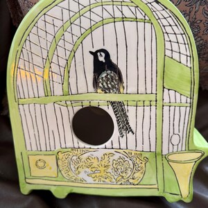 May include: A ceramic birdhouse shaped like a cage, painted in white and light green. A black and white bird is perched inside. The birdhouse has a round opening and decorative floral accents.
