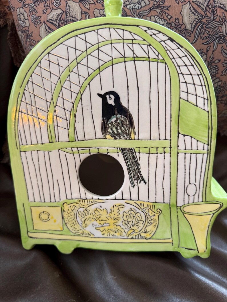 May include: A ceramic birdhouse shaped like a cage, painted in white and light green. A black and white bird is perched inside. The birdhouse has a round opening and decorative floral accents.