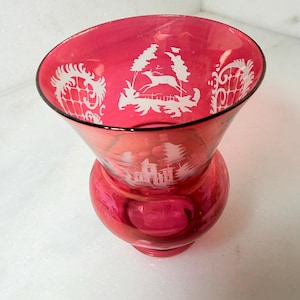 May include: A ruby red glass vase with a flared top and a rounded base. The vase features white etched designs of a deer, trees, and architectural elements. The rim is outlined in black.
