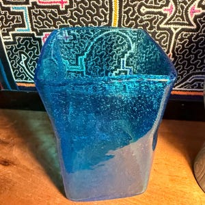 Peut inclure: Un vase en verre bleu avec une forme géométrique unique. Le vase présente un effet de dégradé, passant d'un bleu profond en haut à une teinte plus claire, presque transparente en bas. Le vase est posé sur une surface en bois.