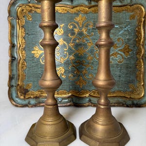 May include: Two antique brass candlesticks with hexagonal bases and ornate, detailed stems. The candlesticks are a warm gold color, set against a teal and gold decorative tray.