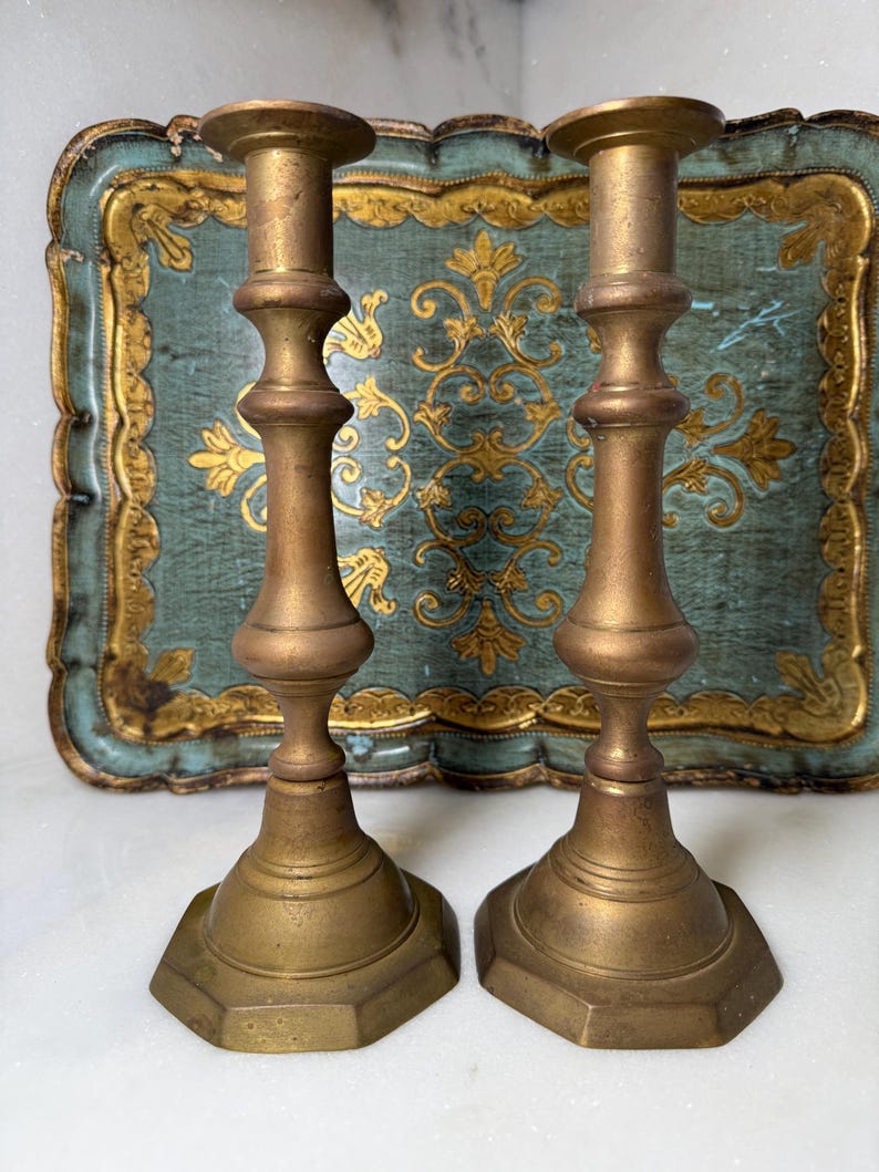May include: Two antique brass candlesticks with hexagonal bases and ornate, detailed stems. The candlesticks are a warm gold color, set against a teal and gold decorative tray.