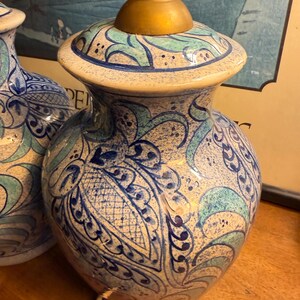May include: A pair of ceramic table lamps with a cream-colored base and intricate blue and teal floral patterns. The lamps have a gold-colored neck and a matching patterned top. The lamps are on a wooden surface.