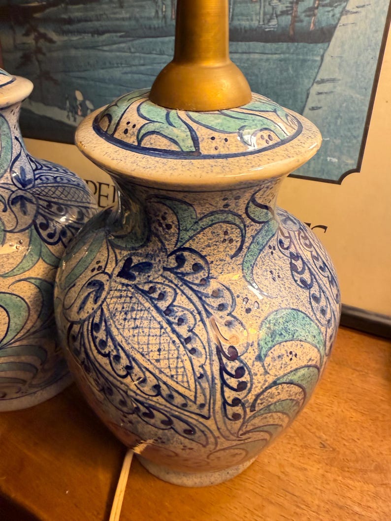 May include: A pair of ceramic table lamps with a cream-colored base and intricate blue and teal floral patterns. The lamps have a gold-colored neck and a matching patterned top. The lamps are on a wooden surface.