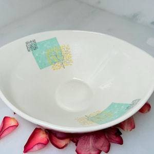 May include: A white, oval ceramic bowl with a decorative design. The bowl has a light blue and yellow tree design on a square background. Red rose petals surround the bowl.