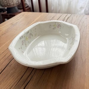 May include: A white, rectangular ceramic dish with gently curved sides. Delicate floral patterns in pink, yellow, and green adorn the rim. The dish sits on a wooden surface, suggesting a vintage or antique aesthetic.