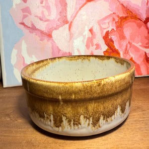 May include: A small, round ceramic bowl with a speckled cream interior and a brown, dripped glaze exterior. The bowl sits on a wooden surface, with a blurred painting of pink flowers in the background.
