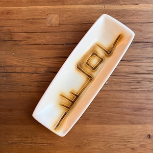 May include: A rectangular ceramic tray with a cream-colored glaze and a dark brown abstract linear design. The tray features smooth, rounded edges and a shallow basin, showcasing a contrasting hand-painted pattern that runs along the center.