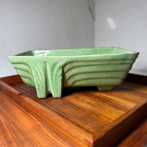 May include: A rectangular, light green ceramic planter with a textured, ridged design. The planter has a unique, stylized leg and is set on a wooden tray. The planter is ideal for small plants or succulents.