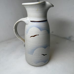 May include: A ceramic pitcher with a light blue and white cloud design and two brown bird silhouettes. The pitcher has a curved handle and a spout for pouring. The pitcher is approximately 9 inches tall.