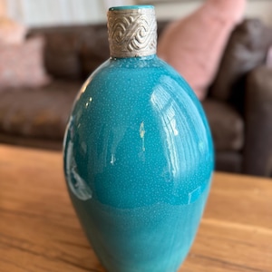 May include: A teal ceramic vase with a speckled finish and a silver-colored metal collar featuring an embossed wave pattern around the neck. The vase has a rounded, bulbous body that tapers toward the top opening, creating a smooth silhouette.