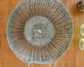 May include: A round, silver-toned wire basket with two arched handles. The basket has a woven design and sits on a wooden surface. The basket's diameter is approximately 14 inches. Other glassware is visible in the background.