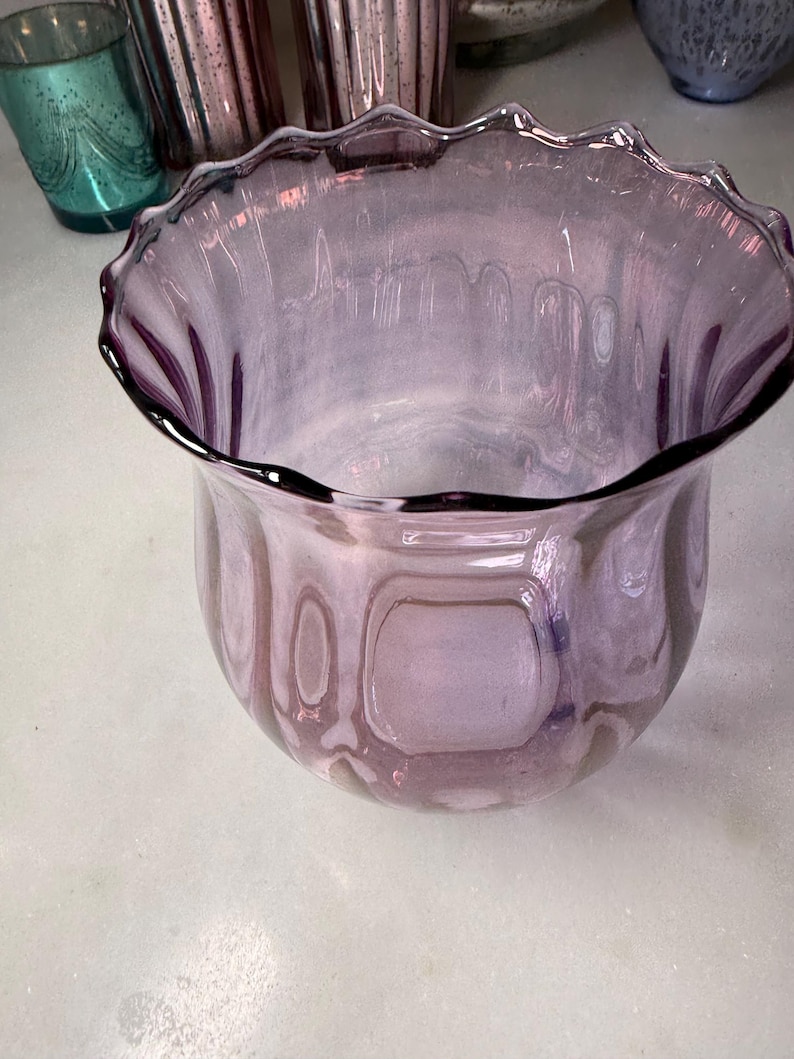 Peut inclure: Un vase d&eacute;coratif en verre violet clair avec un bord festonn&eacute;. Le vase a un corps arrondi et une ouverture l&eacute;g&egrave;rement &eacute;vas&eacute;e. Le verre a un aspect textur&eacute; et le bord est d'une teinte violette plus fonc&eacute;e.