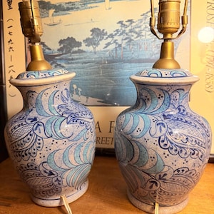 May include: Pair of ceramic table lamps with blue and white floral patterns. The lamps have a vase-like shape with brass fittings and a cream-colored base. The lamps are connected to a power source with a white cord. The background features a framed print.