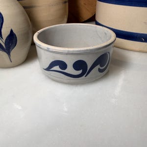 May include: A round, light gray stoneware bowl featuring a hand-painted cobalt blue swirl design around the exterior. The bowl has a smooth, glazed finish and a thick rim, showcasing a classic pottery aesthetic with a simple, curved decorative pattern.