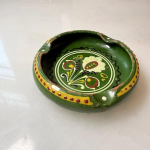 May include: A round green ceramic ashtray featuring a hand-painted floral motif in the center with white, yellow, and brown accents. The rim is decorated with a yellow band containing brown dots, and the piece has two notches for resting items.