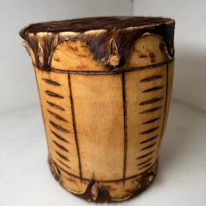 May include: A small, cylindrical drum with a light brown wooden body and a dark brown, fur-covered top. The drum's body features vertical, dark lines and is trimmed with fur at the top and bottom. The drum is likely handmade.