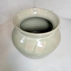 May include: A handmade, light green ceramic vase with a wide, flared rim. The vase has a rounded body and a smooth, glazed finish. The interior is a darker shade of green, and the vase is approximately 8 inches tall.