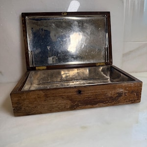 May include: A rectangular wooden box with a hinged lid and a metallic silver-toned interior lining. The exterior wood features a weathered, dark brown finish with visible grain and wear. The box includes two brass-colored hinges and a small keyhole.