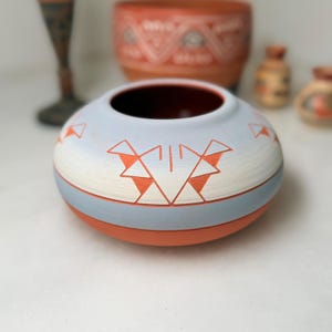 Può includere: Una ciotola in ceramica bassa e arrotondata con finitura opaca, caratterizzata da una sezione superiore azzurra e una base color arancio terracotta. L'esterno è decorato con motivi geometrici triangolari incisi.