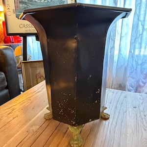 May include: A black, hexagonal planter with a flared top and decorative green claw feet. The planter is made of metal and has a slightly distressed finish, suggesting an antique or vintage aesthetic. The planter is approximately 24 inches tall.