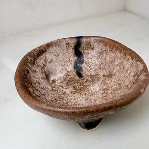 May include: A small, round, footed ceramic bowl with a textured, mottled finish in shades of beige, brown, and cream. The interior features a distinct, dark, vertical brushstroke pattern that extends from the rim toward the center of the bowl.