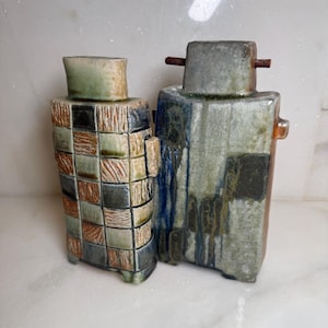 May include: Two rectangular ceramic vases with distinct glazes. The left vase features a grid of textured brown and green squares. The right vase has a mottled grey and blue glaze with small protruding side handles and a top crossbar detail.