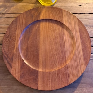 May include: A large, round wooden charger plate with a recessed center. The plate is made of light brown wood with visible grain patterns. Two yellow glass vases are visible in the background, resting on a wooden surface.