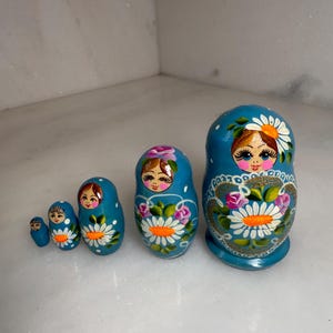 May include: A set of six blue Matryoshka nesting dolls. Each doll features a painted face and floral designs in white, pink, and gold. The largest doll is approximately 5 inches tall, with the smallest being about 1 inch.