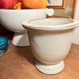 May include: A cream-colored ceramic vase with a pedestal base and a wide rim. The vase has a speckled texture and a slightly flared shape. A white ceramic bowl with fruit is in the background.