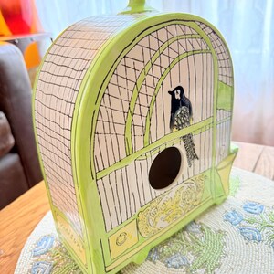 May include: A ceramic birdhouse in the shape of a cage, painted in light green with black and white details. The birdhouse features a black bird inside and decorative yellow accents.