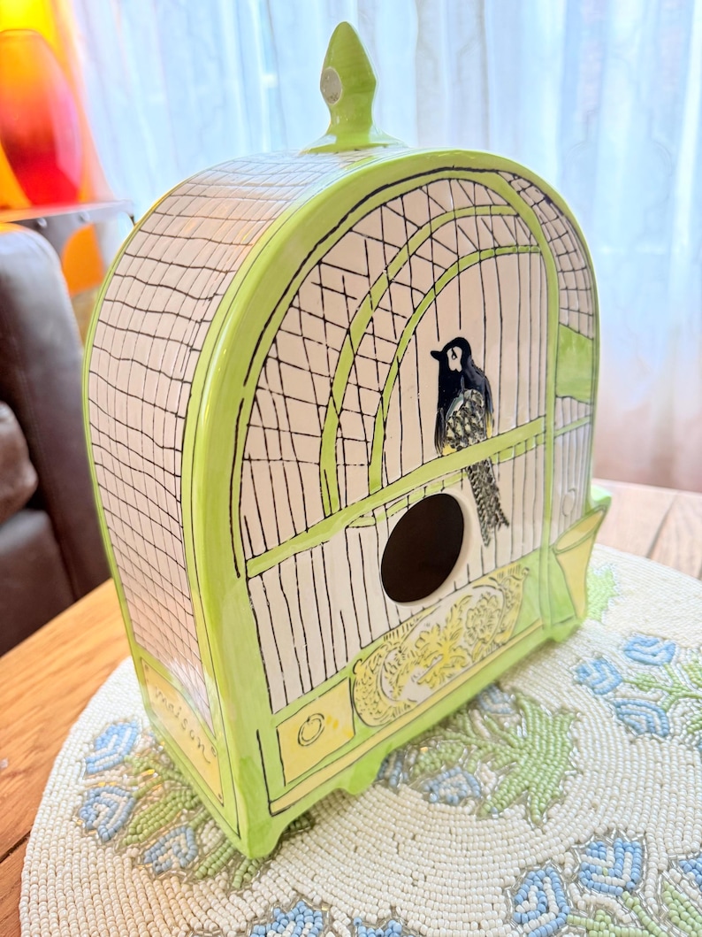 May include: A ceramic birdhouse in the shape of a cage, painted in light green with black and white details. The birdhouse features a black bird inside and decorative yellow accents.