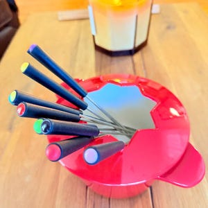 May include: A red fondue pot with black handles and colorful tips, containing fondue forks. A lit candle in a glass jar sits in the background. The fondue pot is on a wooden table.