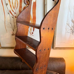 Pode incluir: Uma prateleira de madeira de três níveis com acabamento castanho escuro. A prateleira tem um design único com laterais curvas e volutas decorativas no topo. As prateleiras são suportadas por um painel traseiro com cabeças de pregos visíveis.
