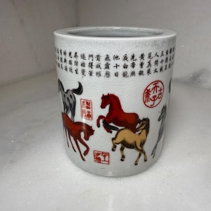 May include: A cylindrical white ceramic brush pot with a crackle glaze finish. The exterior features multiple illustrations of horses in red, brown, and grey, interspersed with red calligraphic stamps and rows of black Chinese characters.