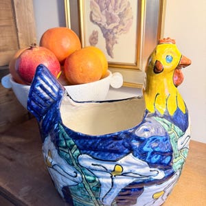 May include: A ceramic hen-shaped planter with a yellow head and blue body, decorated with white floral patterns. The planter is on a wooden surface, with a bowl of oranges and pomegranates in the background.