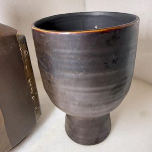 May include: A dark brown ceramic vessel with a wide, rounded body and a pedestal base. The exterior features a subtle horizontal ribbed texture and a smooth, matte finish, while the interior is a deep, solid black with a smooth surface.