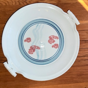 May include: White ceramic serving plate with a blue and pink floral design. The plate features a wide rim and two bamboo-style handles. The center of the plate has a blue ring with pink floral accents and light blue vertical lines.