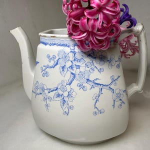 Könnte beinhalten: Weiße Keramik-Teekanne mit blauem Blumenmuster und einem rosa Hyazinthen-Blumenarrangement. Die Teekanne hat einen Ausguss, einen Henkel und einen blauen Rand. Das Blumendesign zeigt Zweige und Blüten.