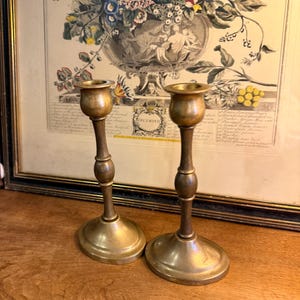 May include: Two matching brass candlesticks with a polished metallic finish. Each candlestick features a wide circular base, a slender stem with a central decorative ring, and a cup-shaped top designed to hold a standard taper candle securely.