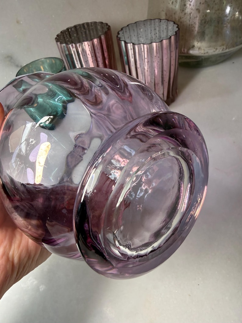 Peut inclure: Un grand vase en verre transparent avec un motif tourbillonnant et marbr&eacute; dans des tons de violet et de blanc. Plusieurs petits photophores en verre rose cannel&eacute; sont en arri&egrave;re-plan. Le vase a une forme arrondie et une base large.