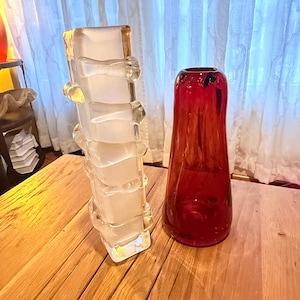 May include: Two decorative glass vases. One is a tall, rectangular vase with a frosted white appearance and clear, rounded accents. The other is a red, teardrop-shaped vase with a glossy finish. Both vases are on a wooden surface.
