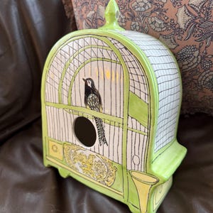 May include: A ceramic birdhouse in the shape of a cage, painted in white, black, and lime green. The birdhouse features a black and white bird perched inside, with a round opening and decorative yellow accents.