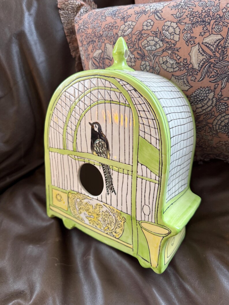 May include: A ceramic birdhouse in the shape of a cage, painted in white, black, and lime green. The birdhouse features a black and white bird perched inside, with a round opening and decorative yellow accents.