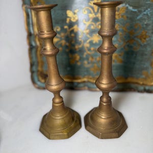 May include: Two antique brass candlesticks with a hexagonal base and a detailed, ornate design. The candlesticks have a warm, golden hue and are set against a blurred background.